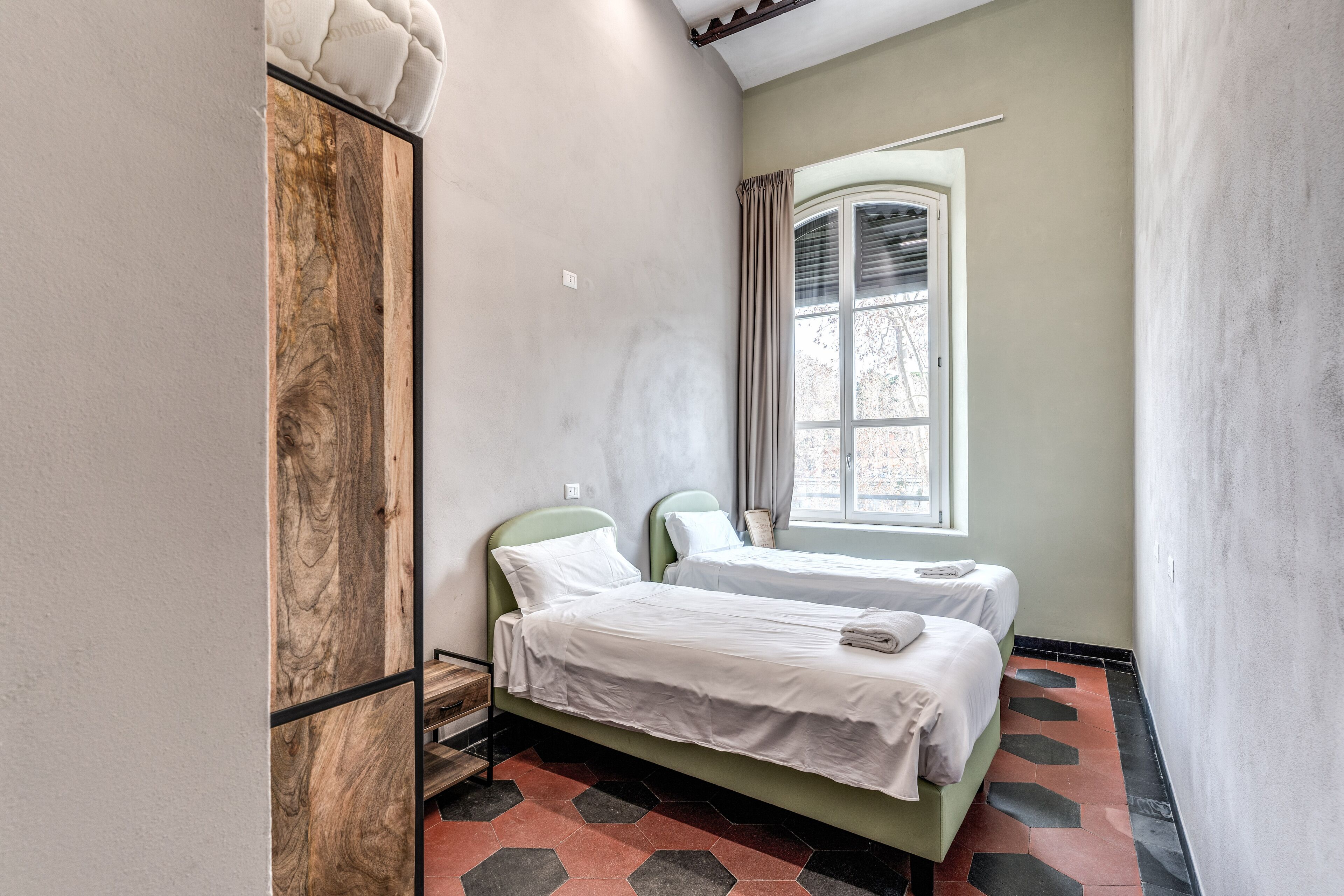 A room with two beds, a window with curtains, a wooden wardrobe, and a hexagonal patterned floor.