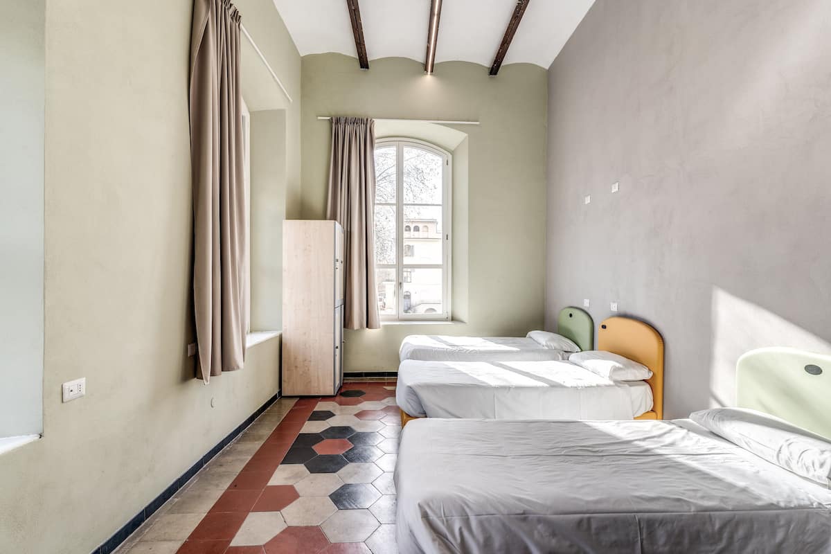 Quarto de hotel com duas camas, um guarda-roupa, uma janela com cortinas e vista para um edifício.