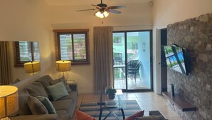 A living room with a sofa, a coffee table, a TV mounted on a stone wall, and a ceiling fan. - Lovely Condo In Playa del Coco! (Coco)