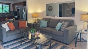 A modern living room with a grey sofa, a glass coffee table, and a kitchen in the background. - Lovely Condo In Playa del Coco! (Coco)