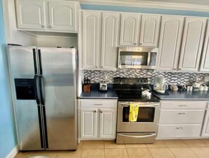 Kitchen area with a stove, microwave, and refrigerator, featuring a tiled backsplash and countertop with a coffee maker and kettle.