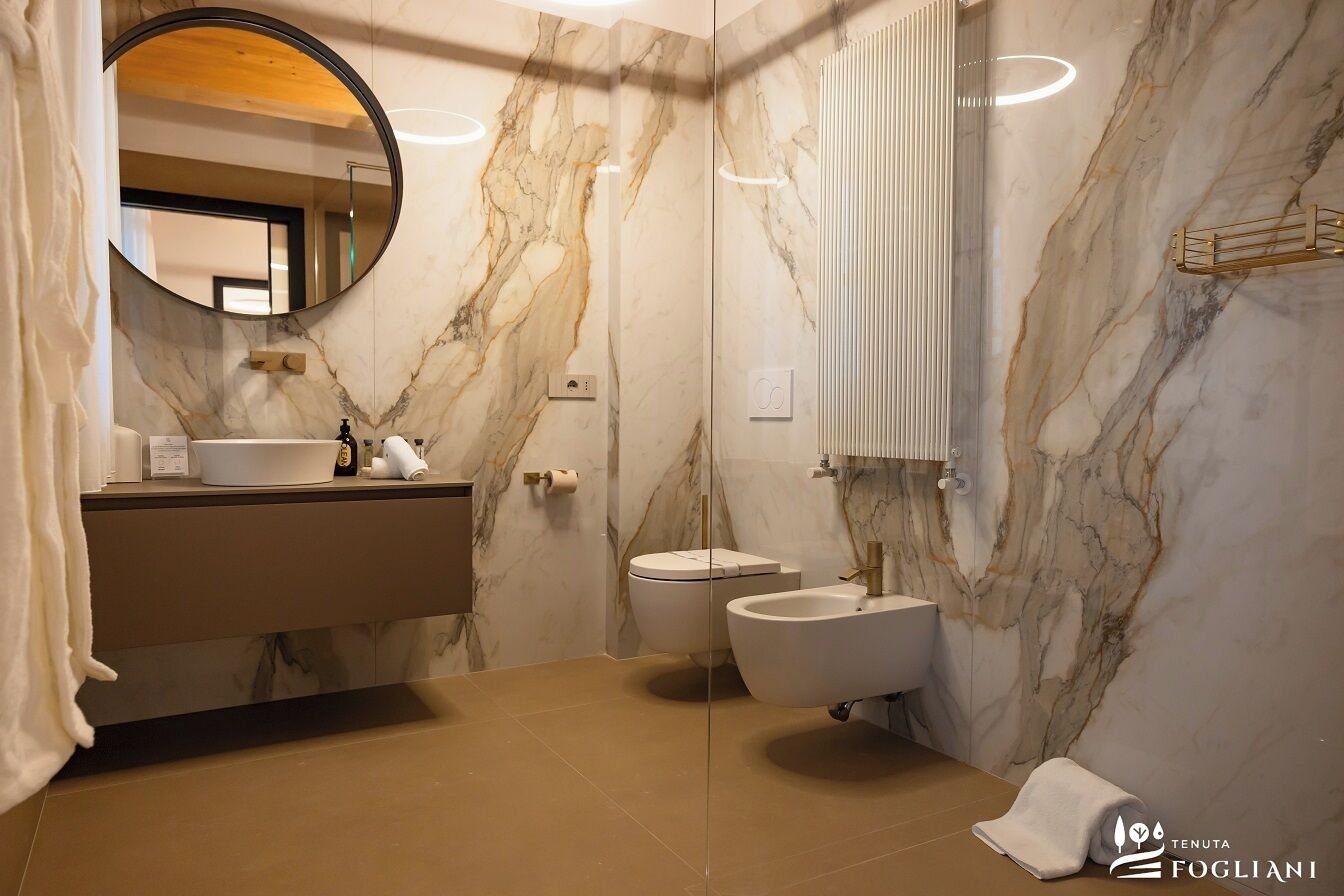 Un baño moderno con pared de mármol, un espejo redondo, un lavamanos, un bidé y una toalla en el suelo.