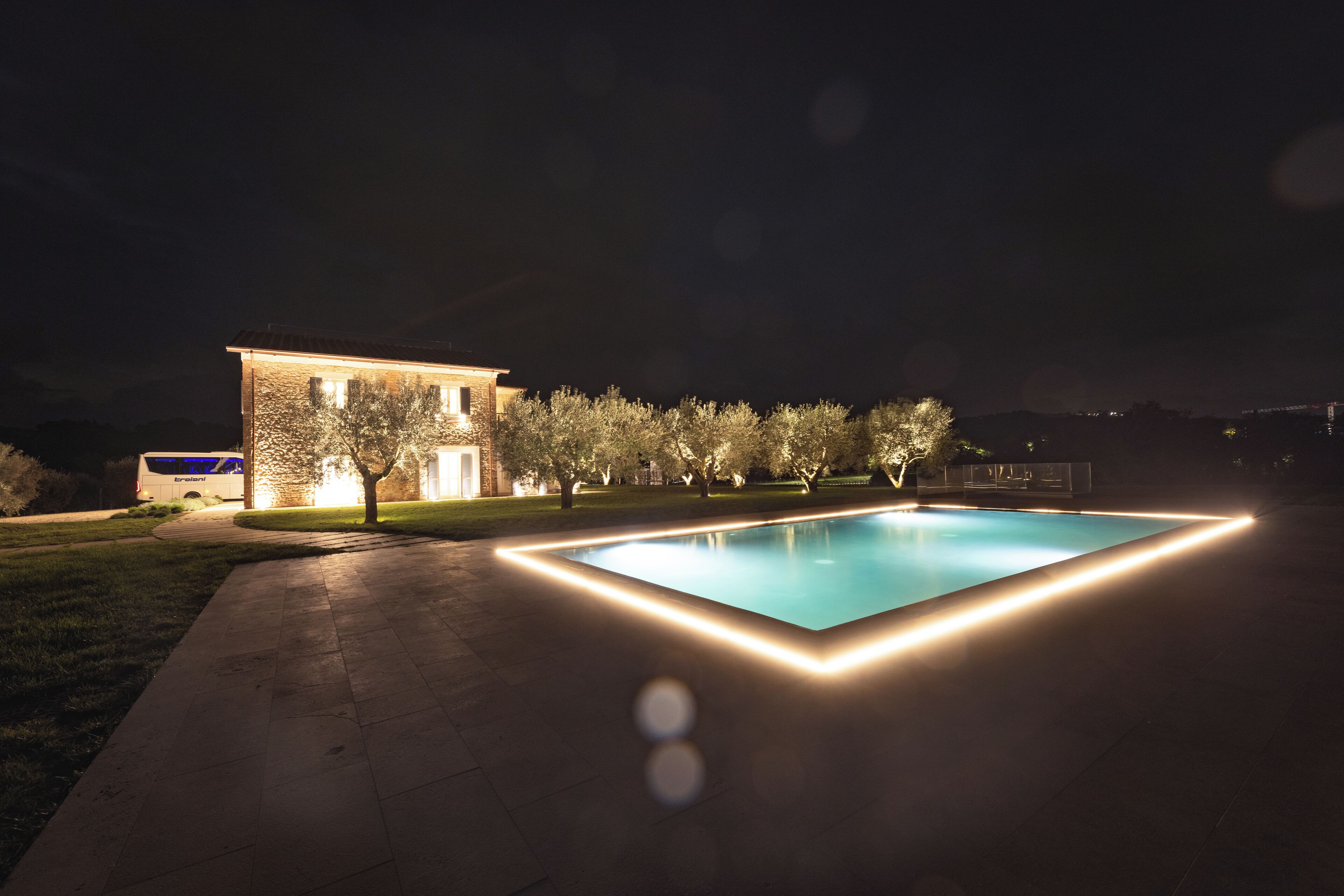 A swimming pool with a light border, a building with illuminated windows, and a bus parked nearby.