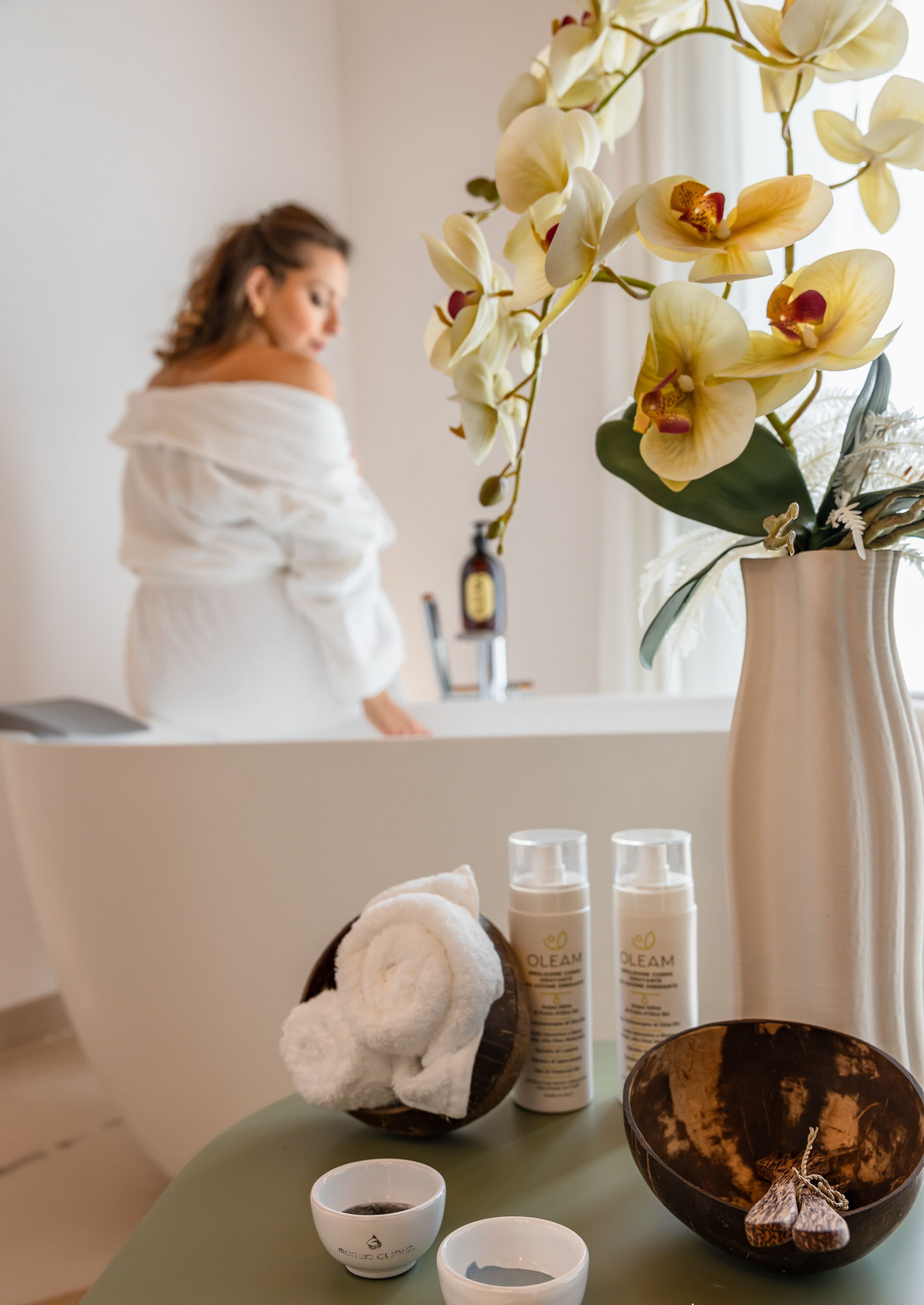 Een spa-achtige omgeving met een witte orchidee vaas, een houten kom met een handdoek en flessen van OLEAM producten op een groene tafel.