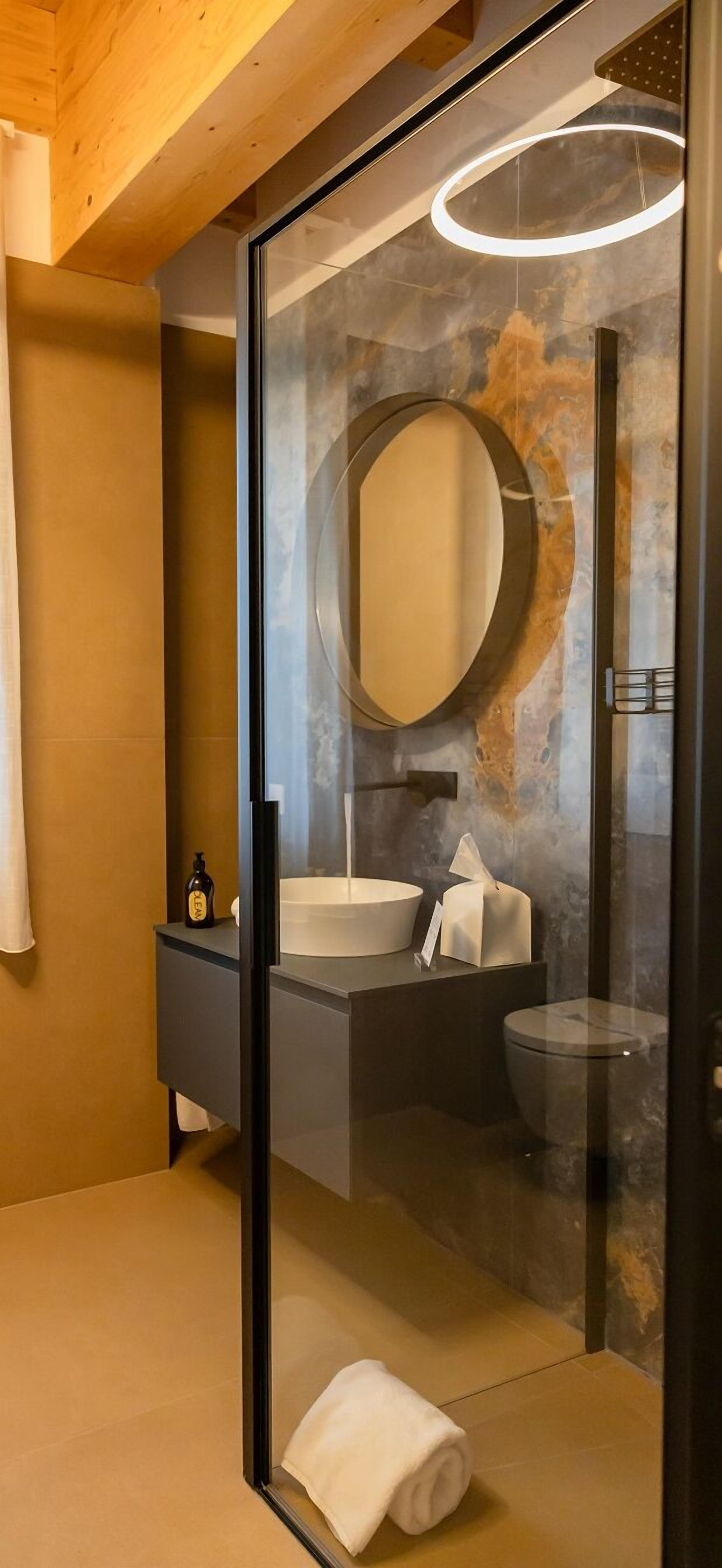 Un baño moderno con ducha envuelta en vidrio, espejo redondo, lavabo blanco y tocador oscuro.