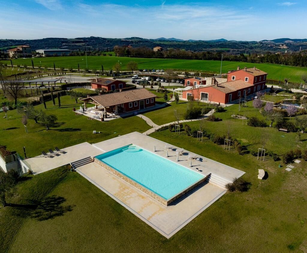 Vista aérea de una propiedad con piscina, un edificio grande de color rojo y campos verdes circundantes.
