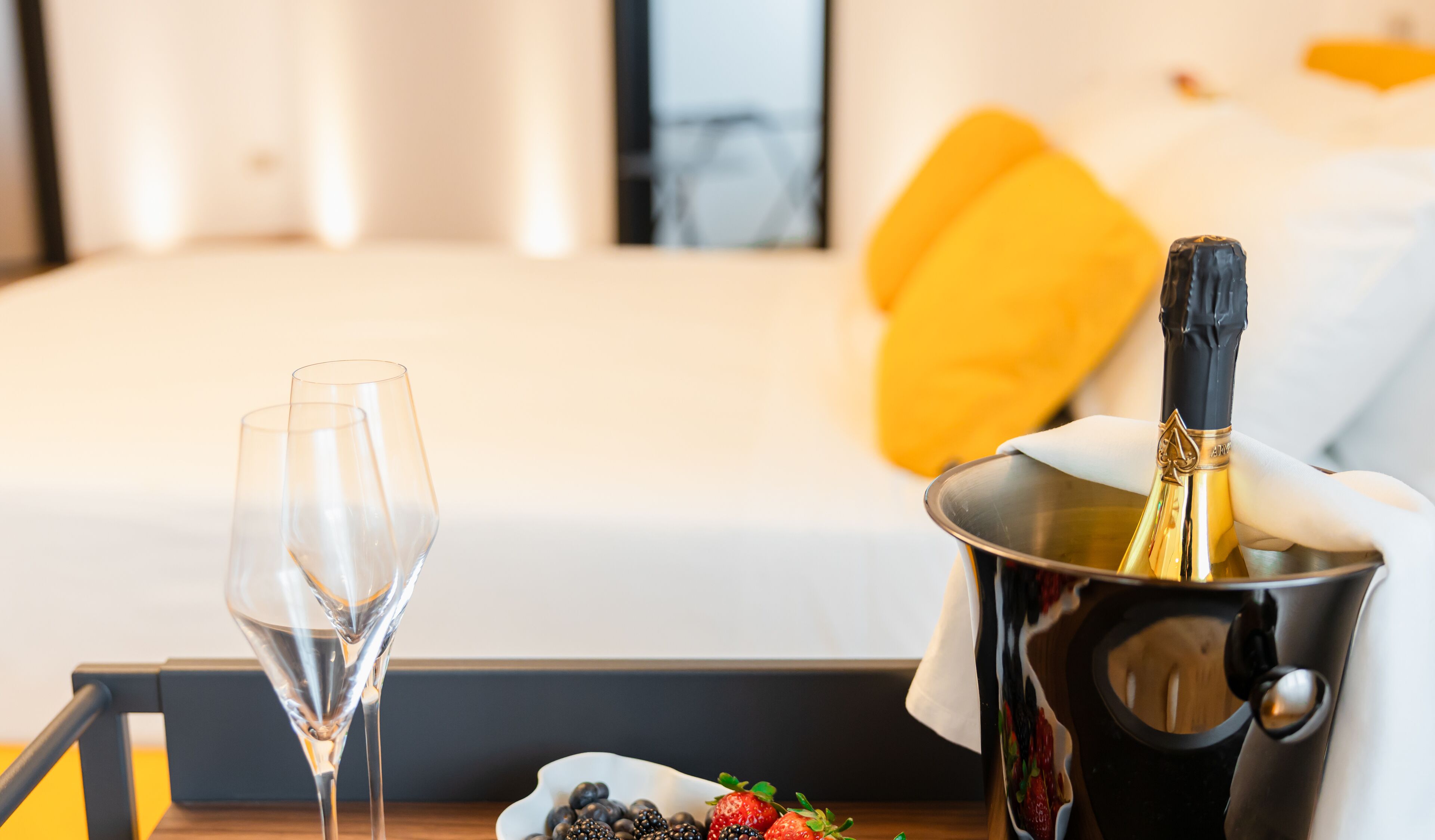 Ein Hotelzimmer mit einem Bett, einem Nachttisch mit Champagnerflasche und Gläsern sowie einer Obstschale.