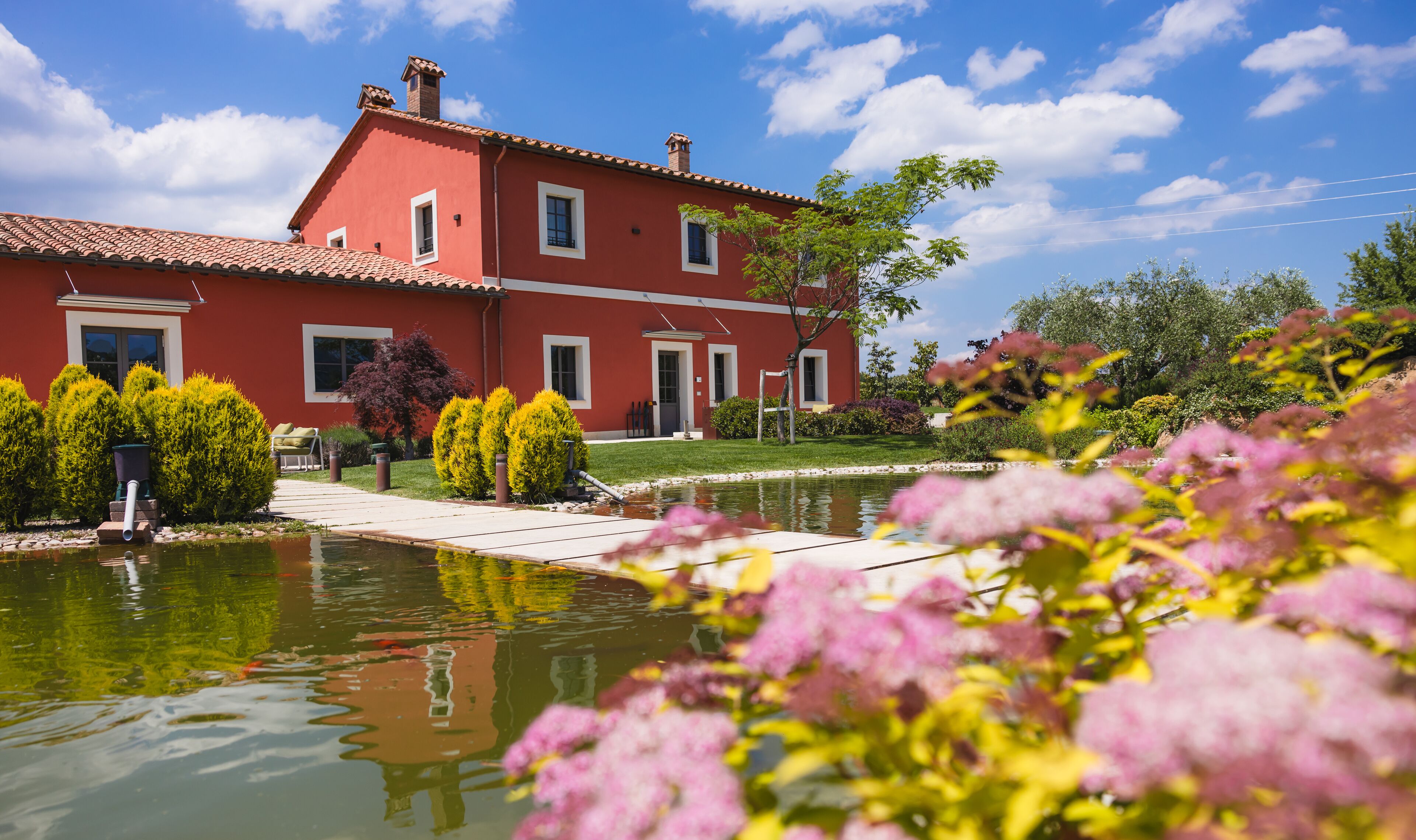 A two-story red building with a tiled roof, a pond in the foreground, and various shrubs and trees around.