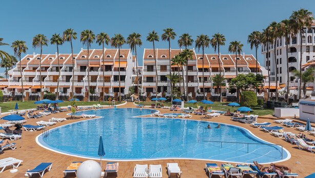 A hotel with a large pool surrounded by sun loungers and umbrellas, palm trees, and a multi-story building with balconies. - GO2TENERIFE Apart’Vera Duplex Santiago 2 (Costa Adeje)