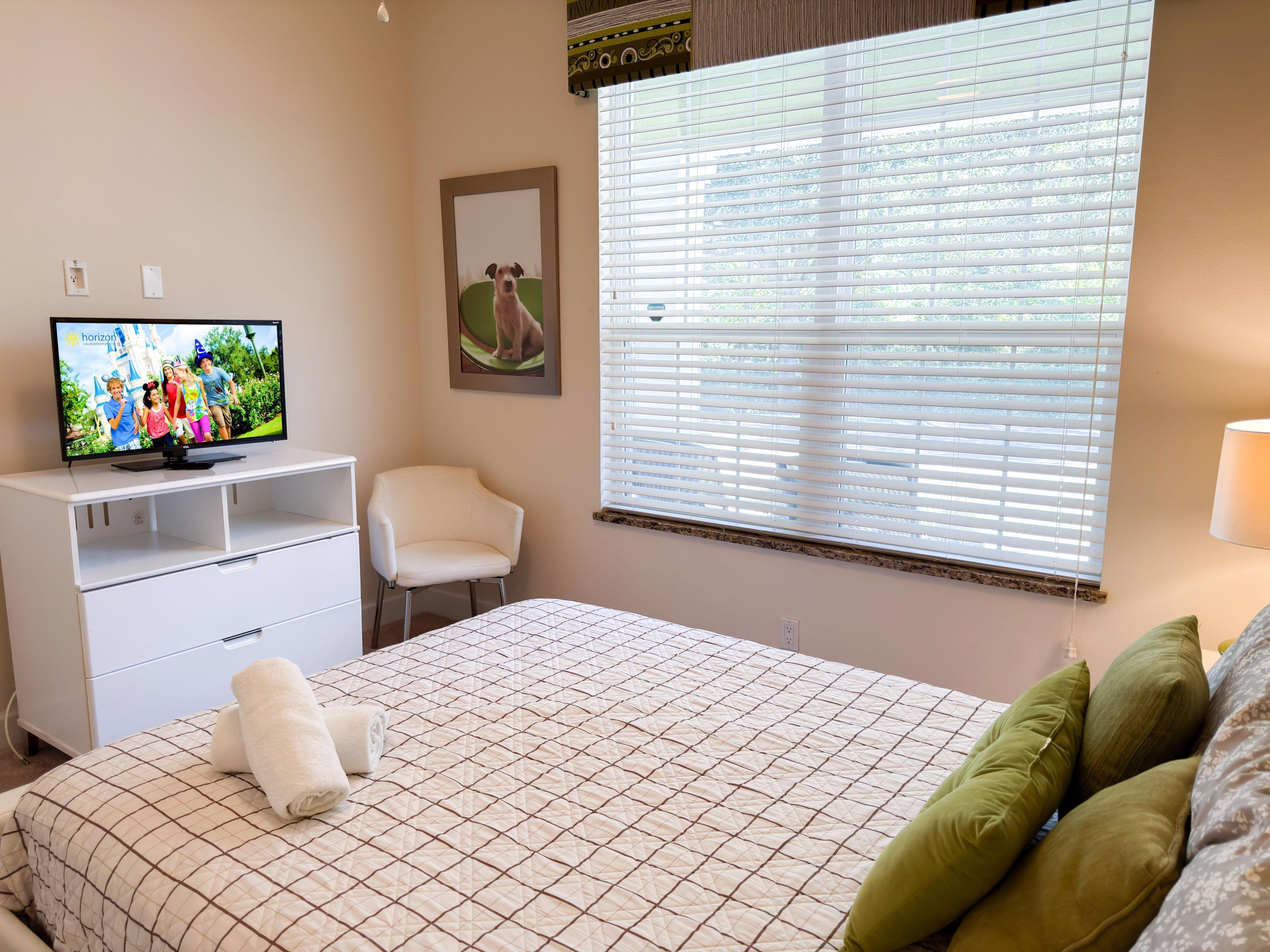 Een slaapkamer met een bed, een televisie op een witte dressoir, een stoel en een raam met jaloezieën.