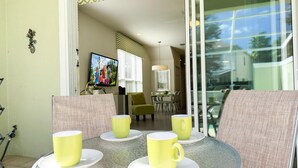 Una mesa redonda de vidrio con tazas y platillos amarillos, un televisor mostrando una caricatura, y un comedor con sillas y una mesa al fondo.