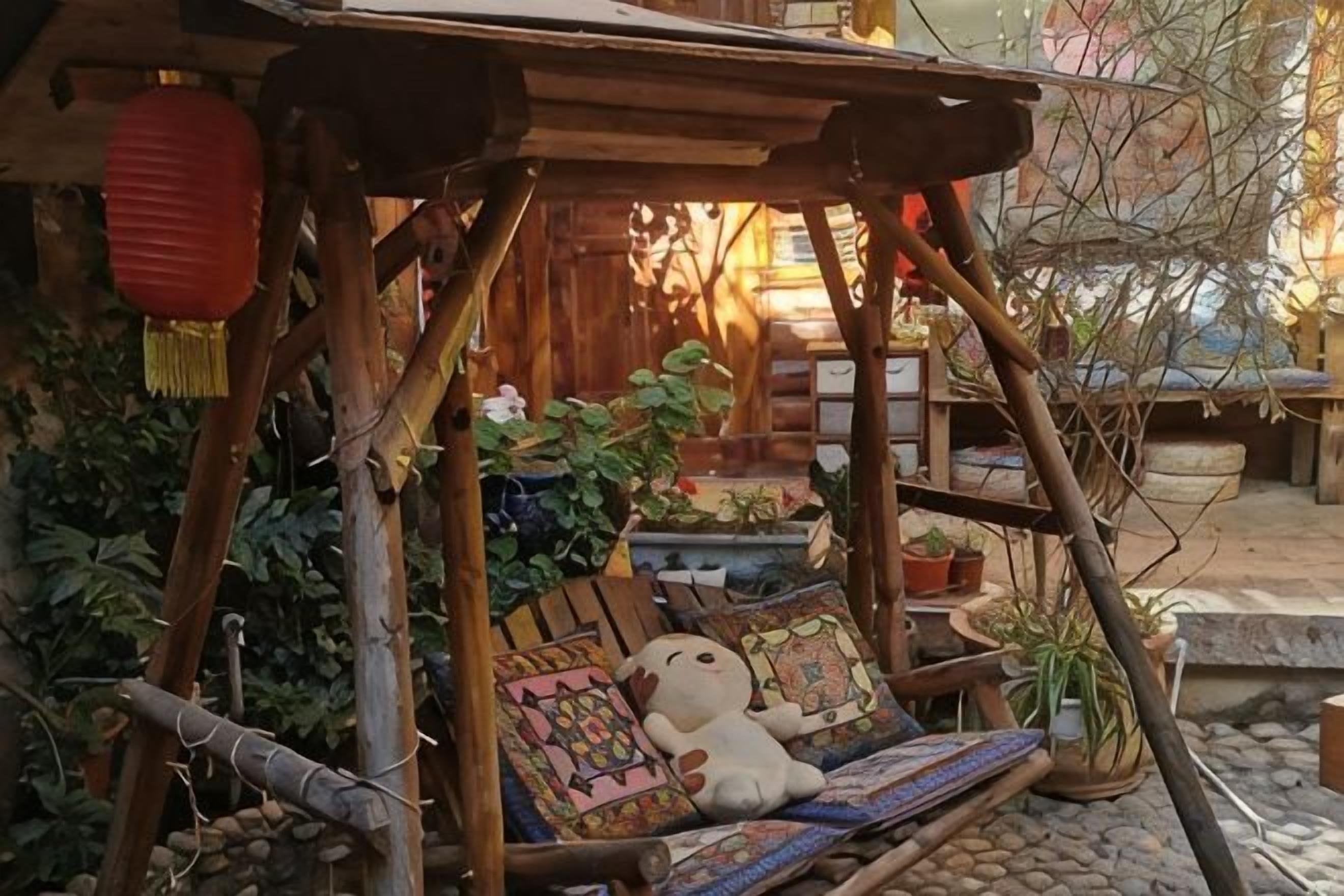 Estrutura de madeira rústica com assento de balanço, rodeada por vasos de plantas e um candeeiro vermelho.