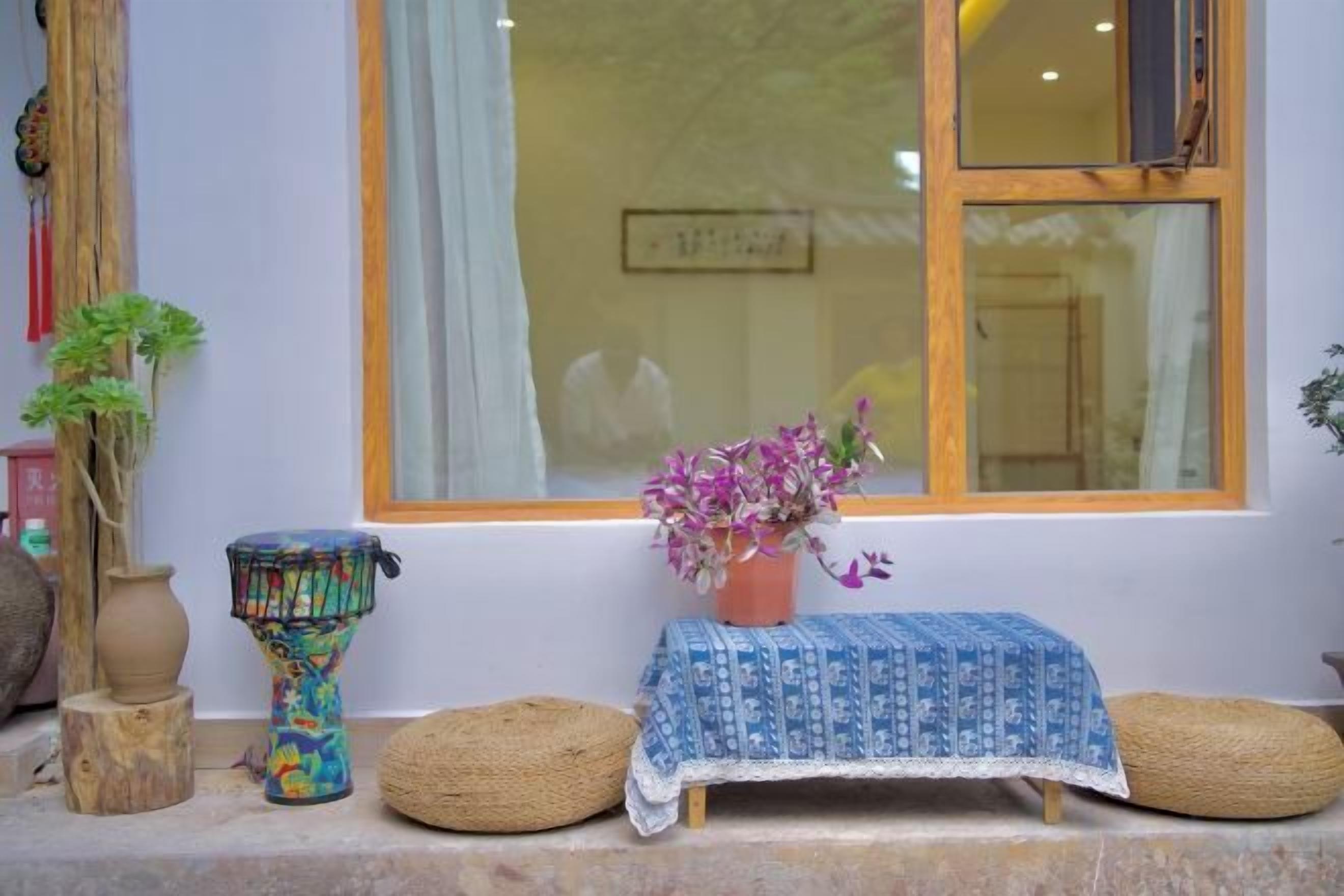 Una pequeña mesa con mantel azul, una planta en maceta y dos taburetes de mimbre frente a una ventana con marco de madera.