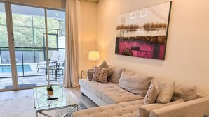 A modern living room with a beige sofa, a glass coffee table, and a view of an outdoor pool area.