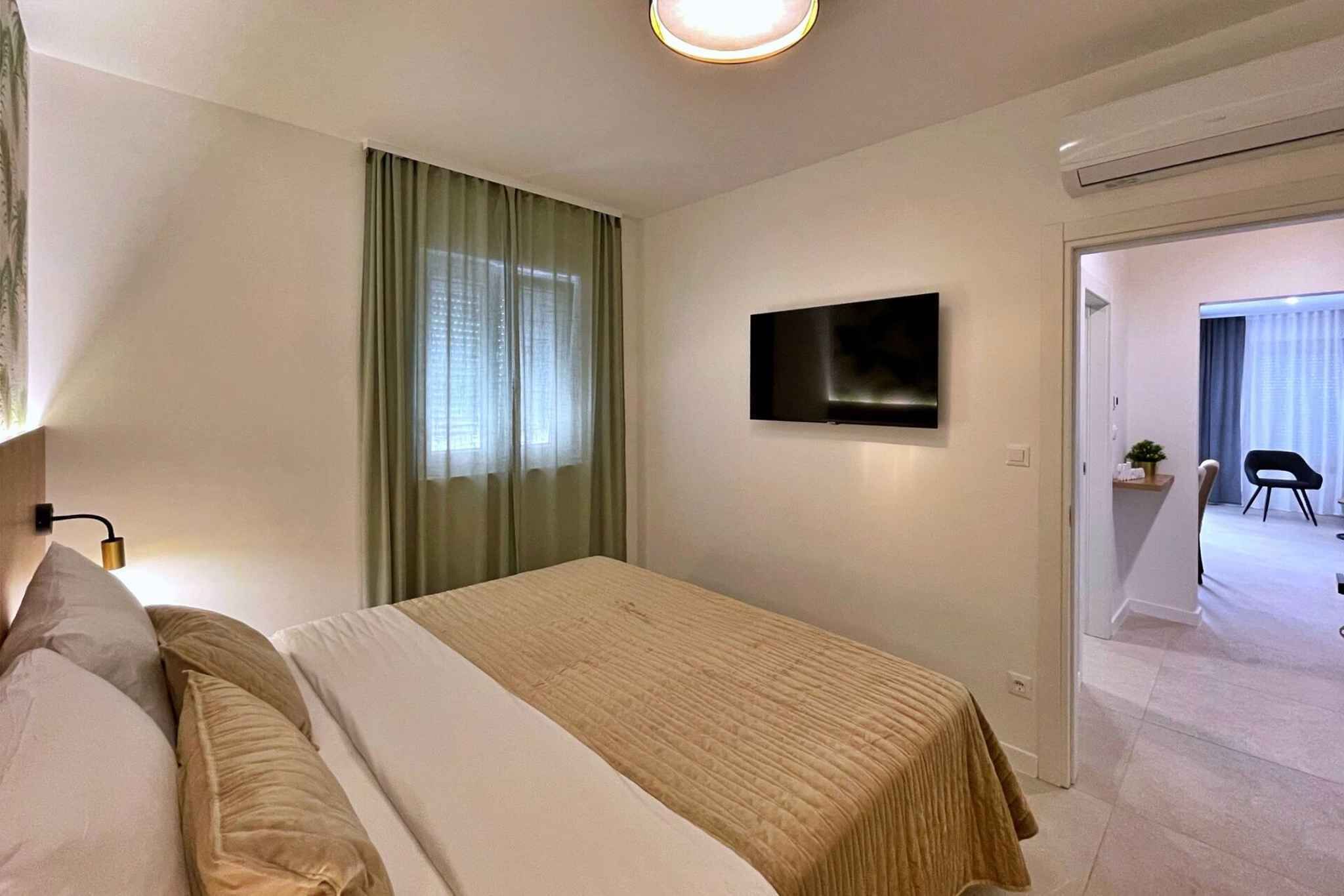 Ein Hotelzimmer mit Bett, flachbildfernseher an der Wand, Fenster mit Vorhängen und einer Tür zu einem anderen Raum.