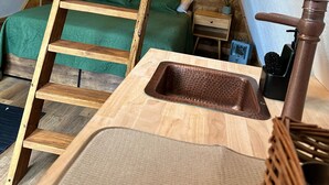 Una stanza compatta e rustica con una scala in legno che conduce a una zona notte soppalcata. Si notano un lavandino e un rubinetto sul pavimento.