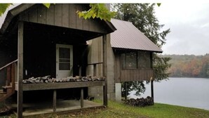 A rustic cabin with a porch and a small deck, overlooking a lake with autumn foliage in the background.