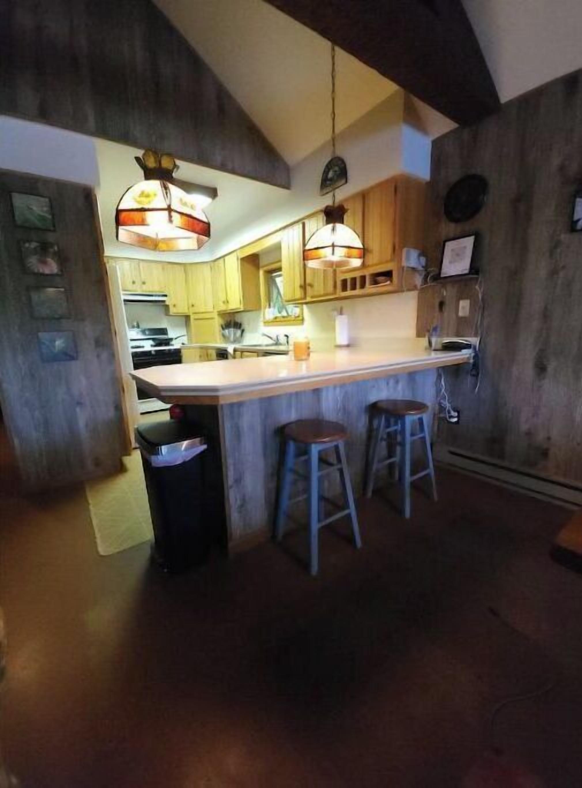 Une cuisine avec des meubles en bois, un îlot central doté de tabourets, un éclairage suspendu et une poubelle visible.