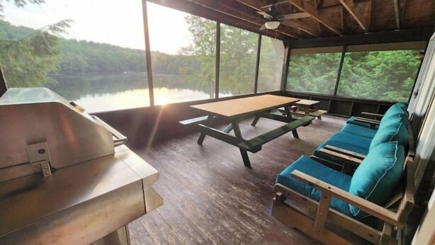 A rustic cabin with a wooden table and benches, a grill, and a view of a lake surrounded by trees.