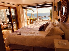 A bedroom with a large bed, a glass door leading to a balcony with a view of the sea, and a small table with a glass bowl.