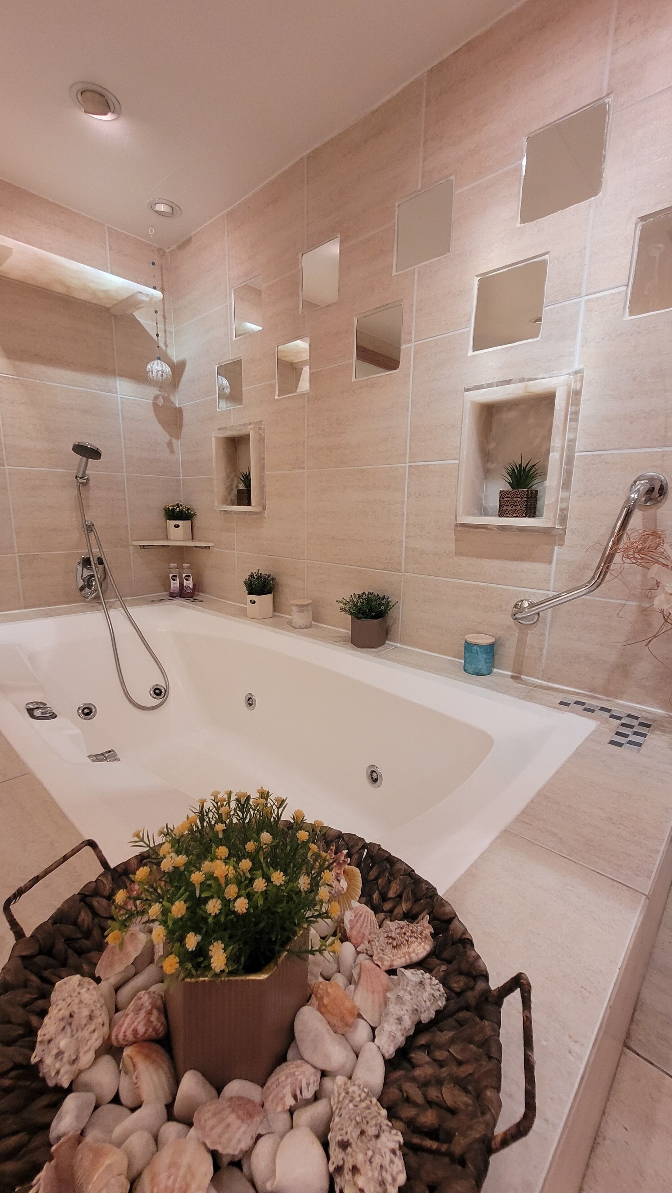 Un baño moderno con una bañera grande, zona de ducha y tocador con espejo. Hay plantas decorativas y un estante pequeño con artículos de aseo.