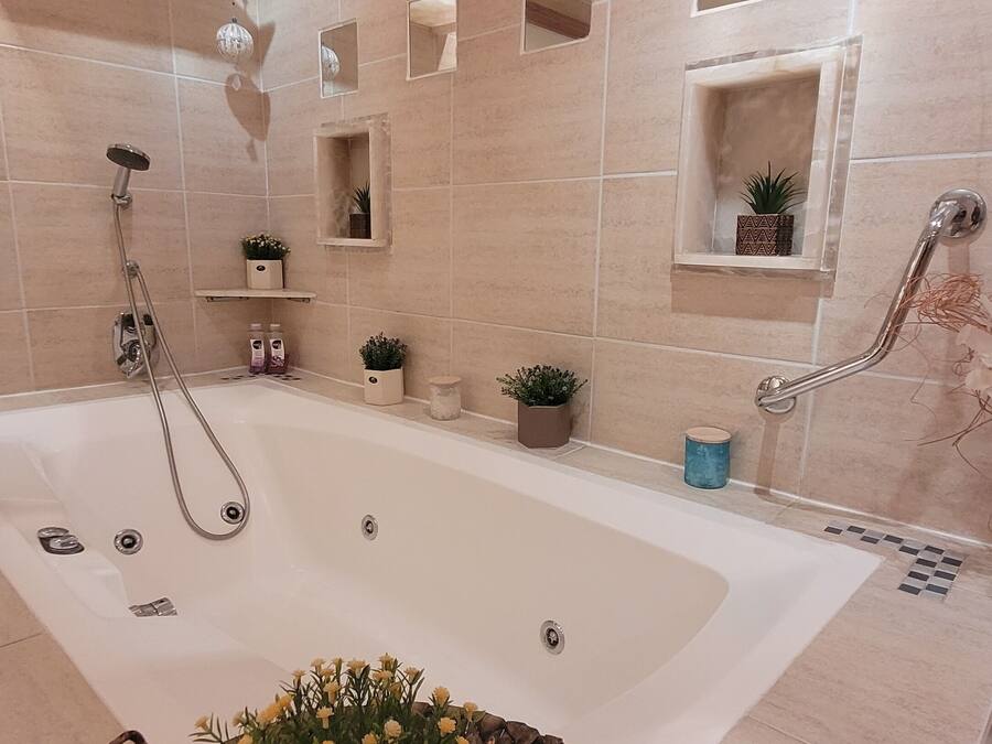 Un baño moderno con una bañera grande, zona de ducha y tocador con espejo. Hay plantas decorativas y un estante pequeño con artículos de aseo.