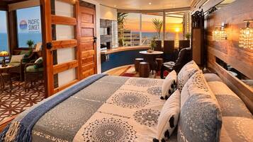 A hotel room with a large bed, a seating area with a view of the sunset, and a wooden door.