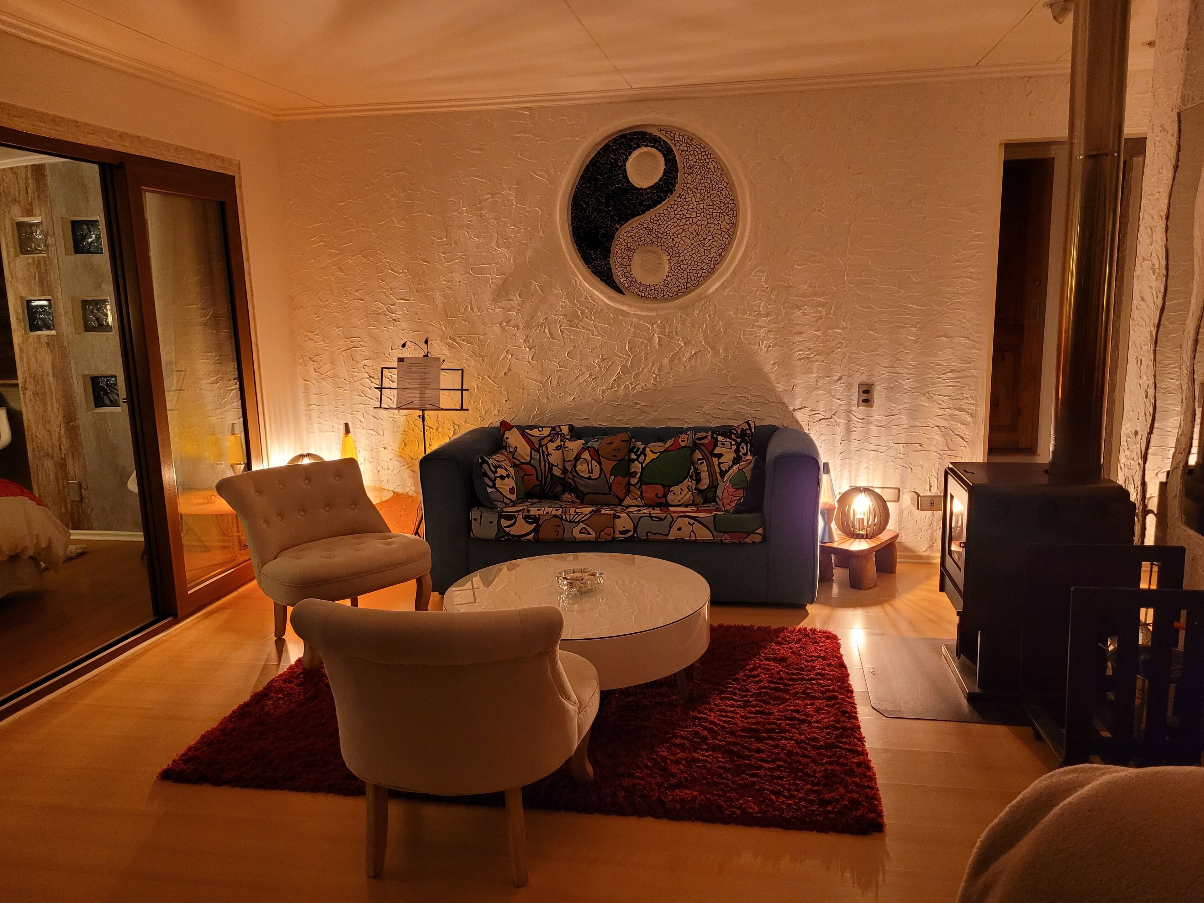 Una sala de estar acogedora con un sofá azul, dos sillones, una mesa redonda de centro y un tapete rojo. Un gran símbolo de yin-yang adorna la pared.