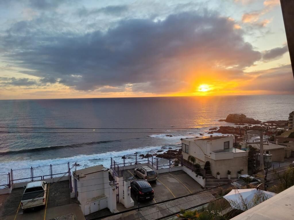 A coastal view with a sunset, rocky shoreline, and a parking area with cars.