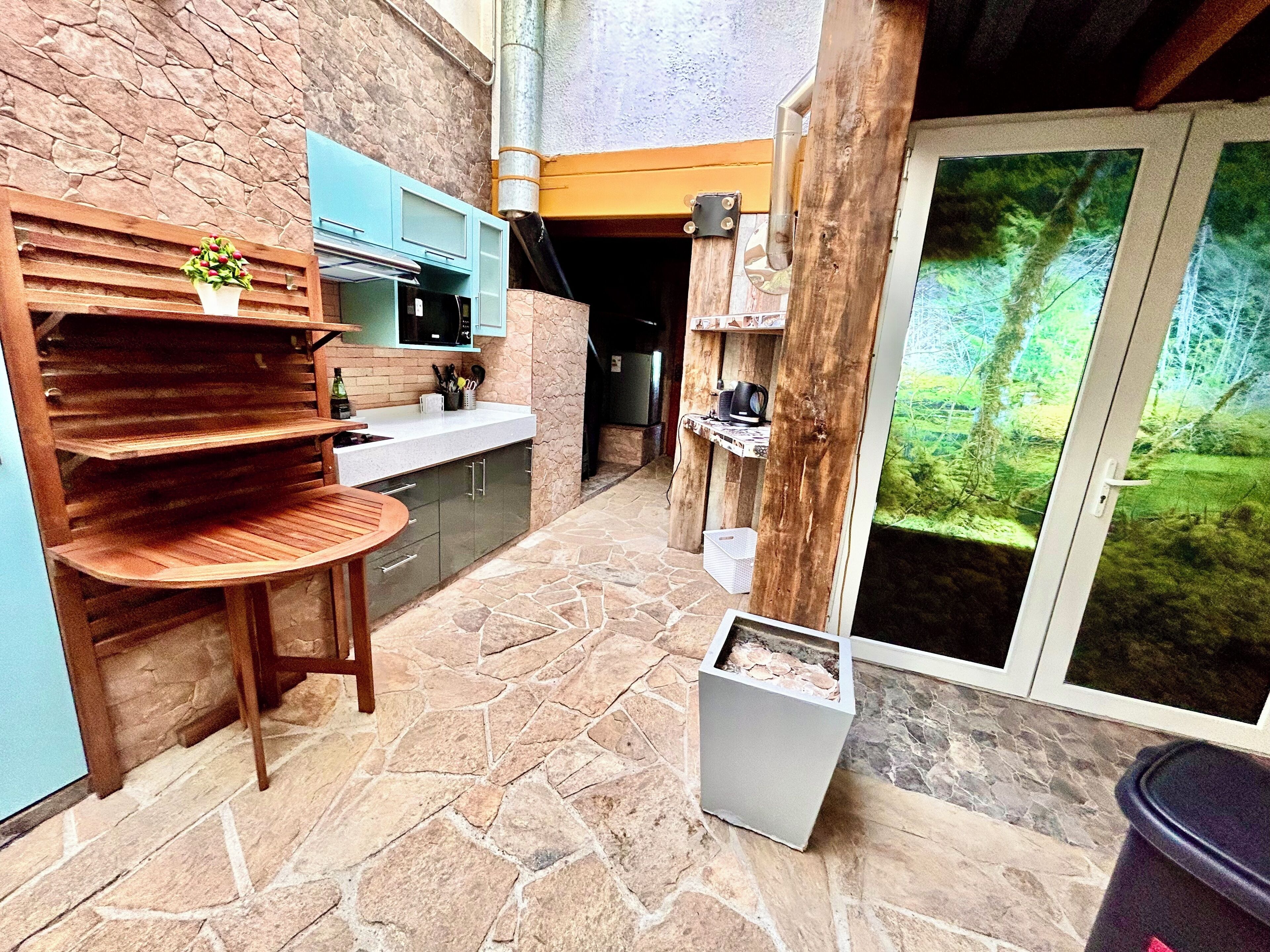 A kitchen with stone flooring, wooden shelves, and a view of greenery through a sliding door.