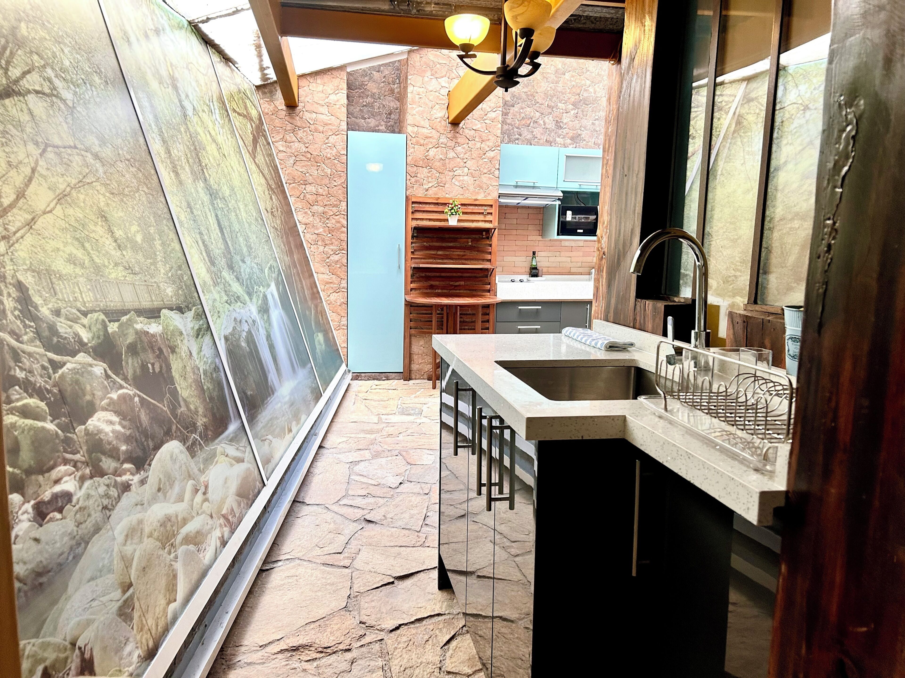 A narrow hallway with a stone floor, a large framed picture on the left, a kitchen area with a sink and bar stools, and a wooden ceiling.