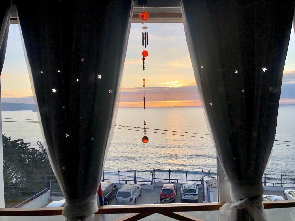 Vista a través de una ventana con cortinas, mostrando un atardecer sobre el océano, un adorno colgante y coches estacionados.