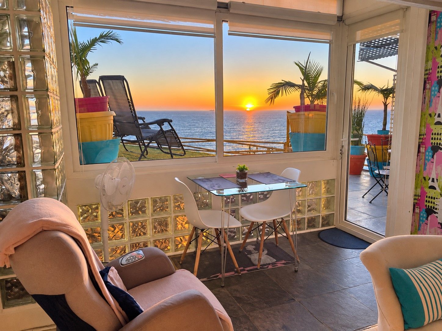 Une chambre avec vue sur le coucher de soleil sur la mer, une table avec des chaises, un fauteuil de massage et un ventilateur.