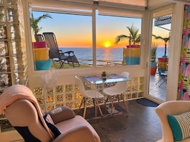 A room with a view of the sunset over the sea, a table with chairs, a massage chair, and a fan.