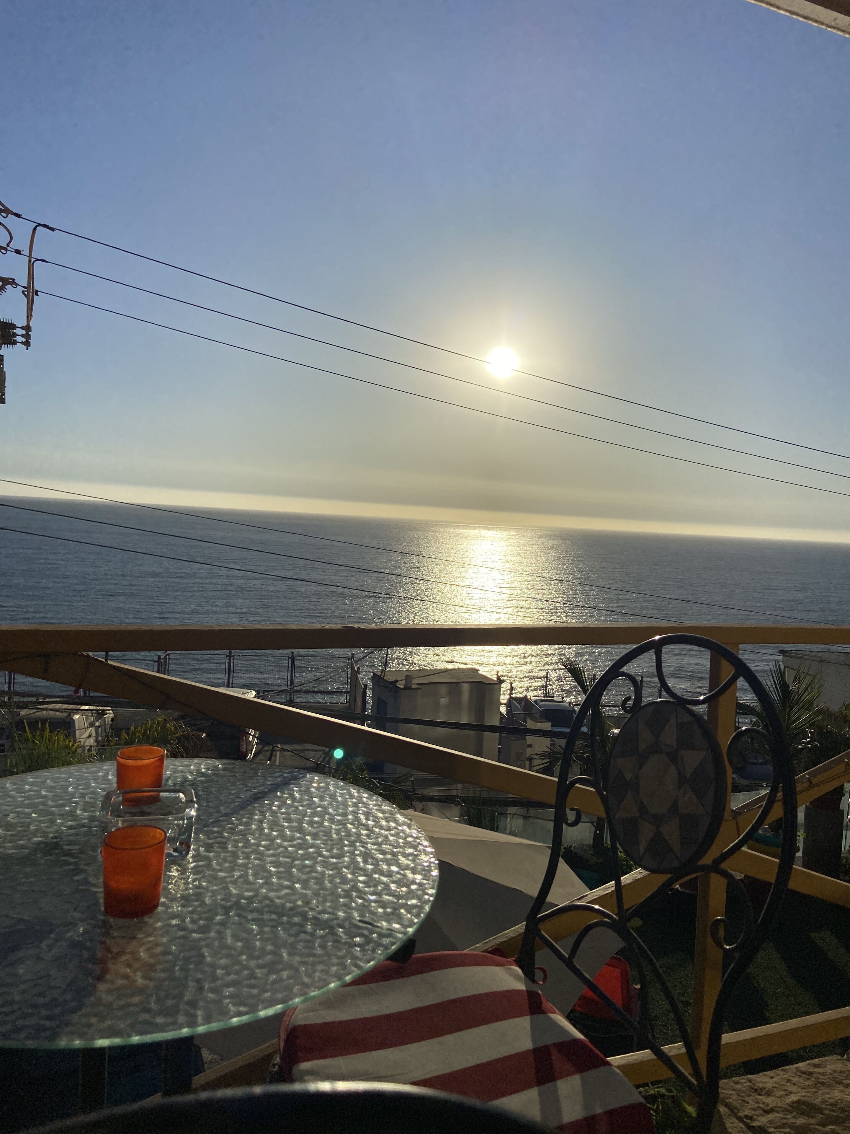 A table with two orange glasses, a patterned cushion, and a view of the ocean with the sun setting or rising.