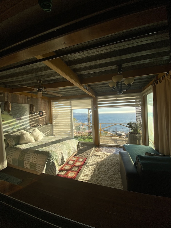 Une chambre avec un grand lit, un plus petit lit et un canapé. On peut voir l’océan et un bâtiment au loin.