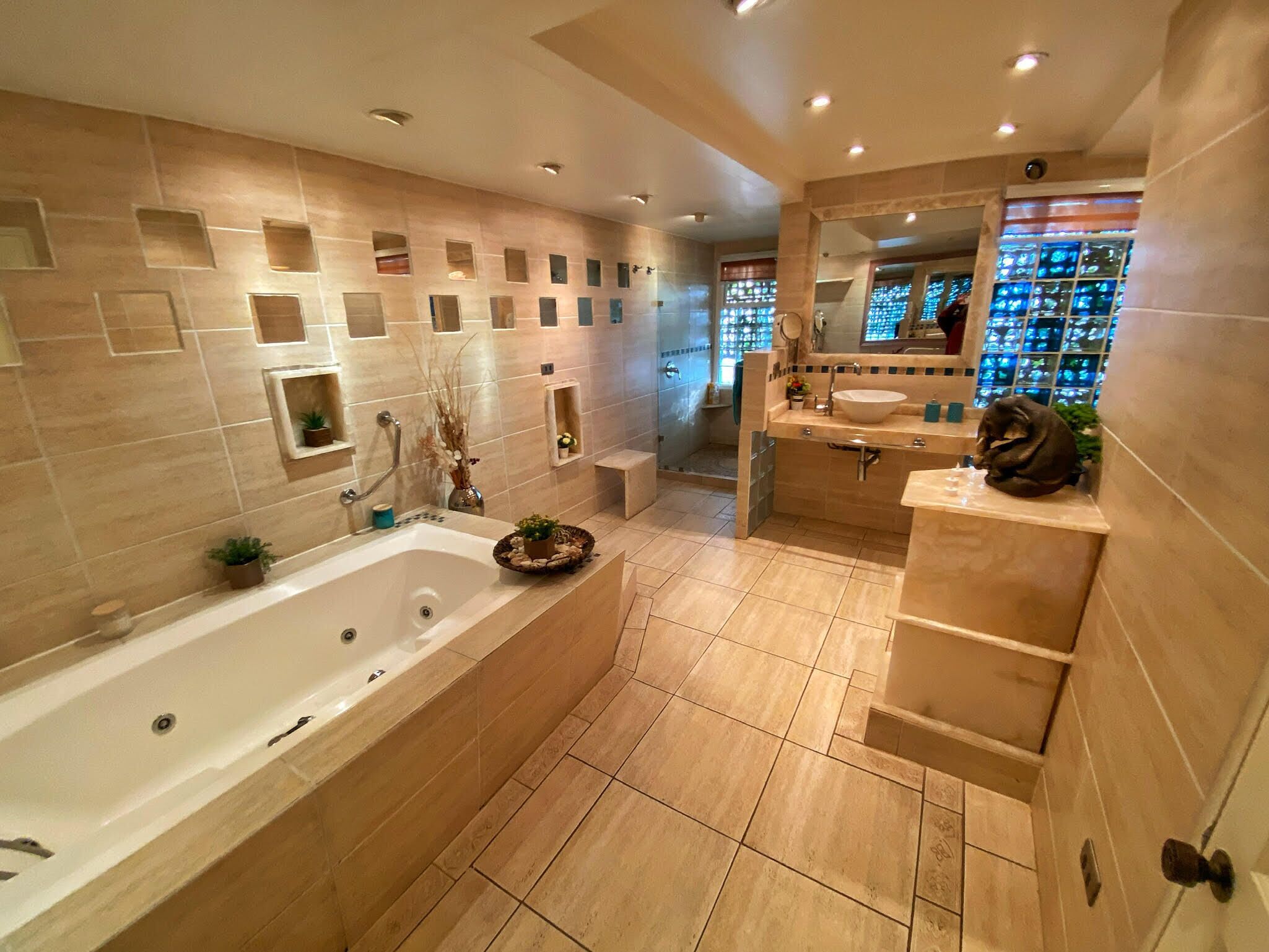 A modern bathroom with a large bathtub, a vanity with a sink, and a mirror. There are multiple shelves with toiletries and a decorative plant on the vanity.