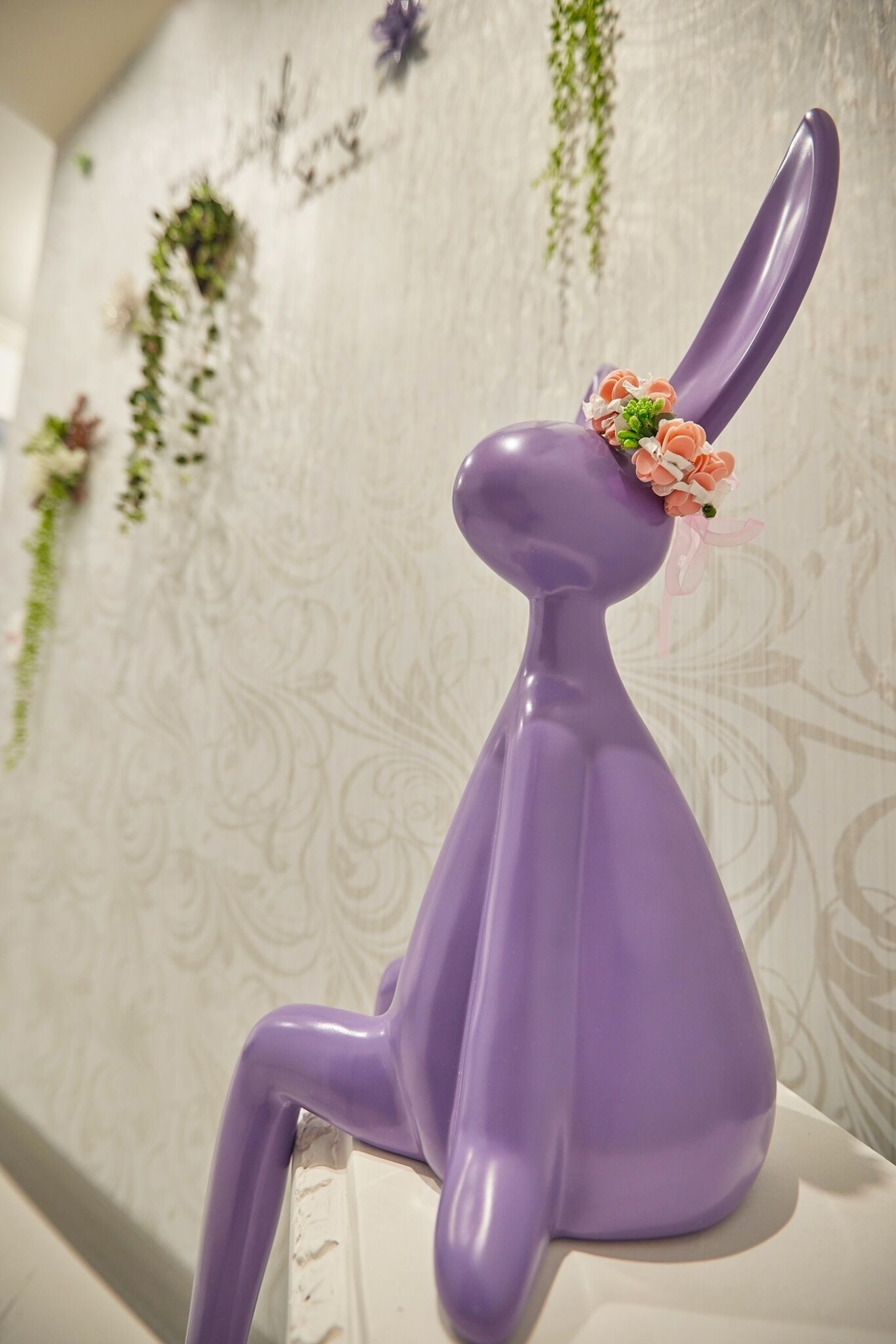 Una scultura di coniglio viola in ceramica con un'decorazione floreale sulla testa, posizionata su una superficie bianca di fronte a un muro decorato.