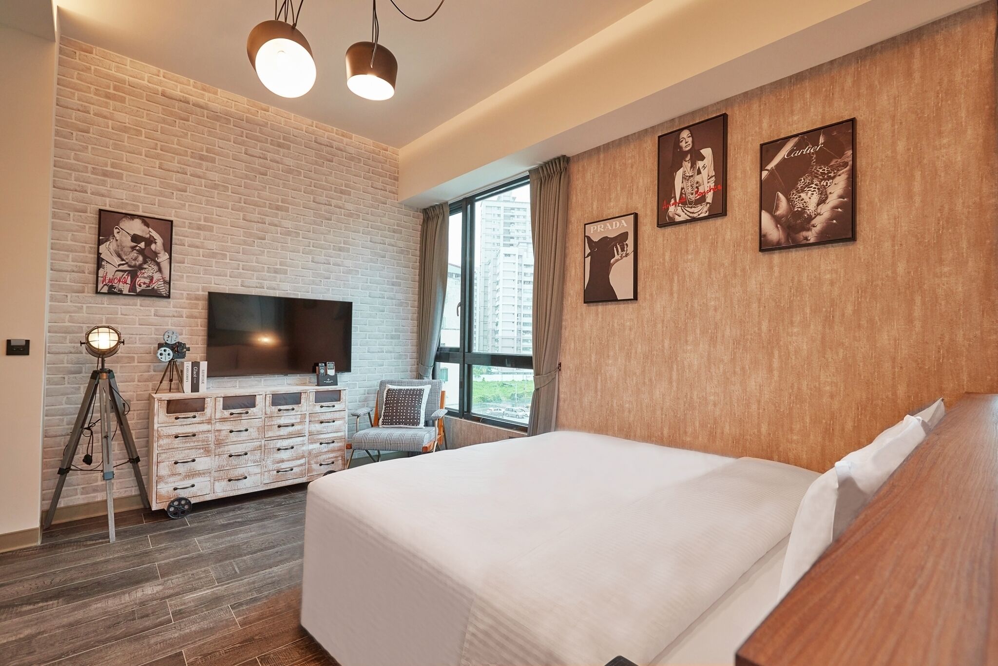 A modern hotel room with a large bed, a flat-screen TV, a wooden dresser, and a brick wall.