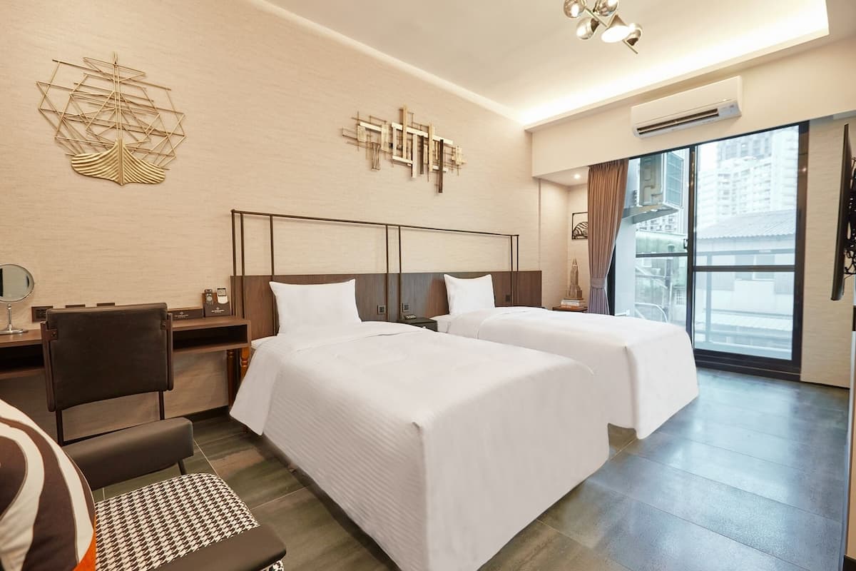 A hotel room with two beds, a desk, a chair, and a large window offering a city view.