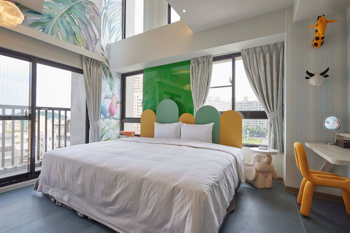 A modern hotel room with a large bed, a desk with a chair, and a view of the city through large windows.