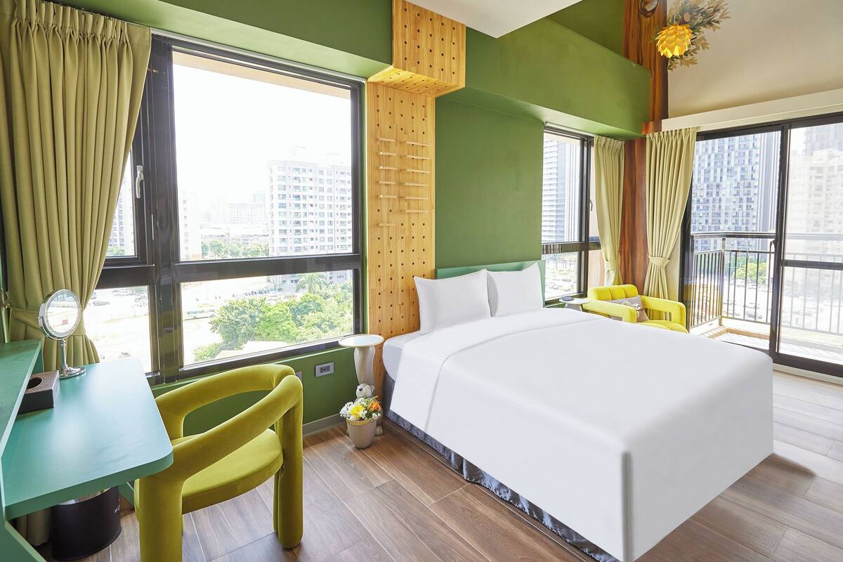 A hotel room with a large bed, a desk, a chair, and a window with a view of buildings.