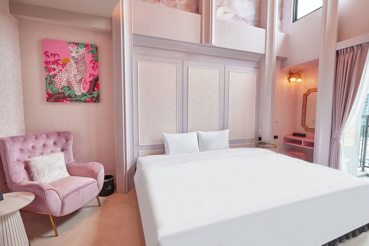 A bedroom with a large bed, a pink armchair, a painting on the wall, and a staircase leading to an upper level.