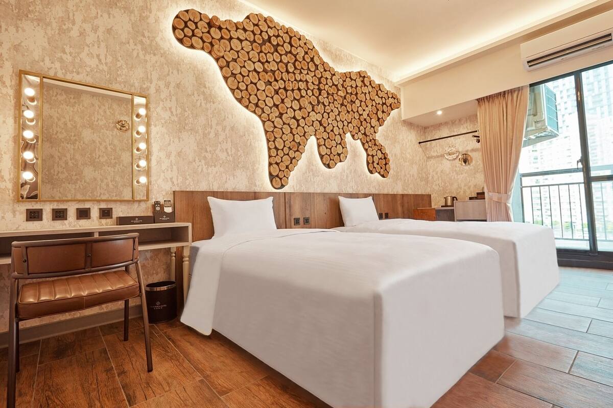 A hotel room with a large mirror with lights, a wooden wall art piece, two beds, a desk, and a chair.