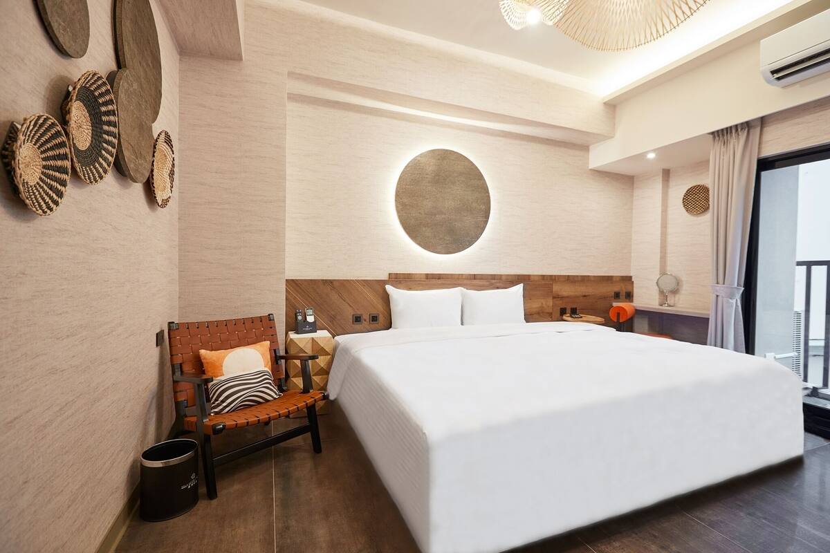 A modern hotel room with a large bed, a wooden headboard, a small seating area, and a circular wall feature.