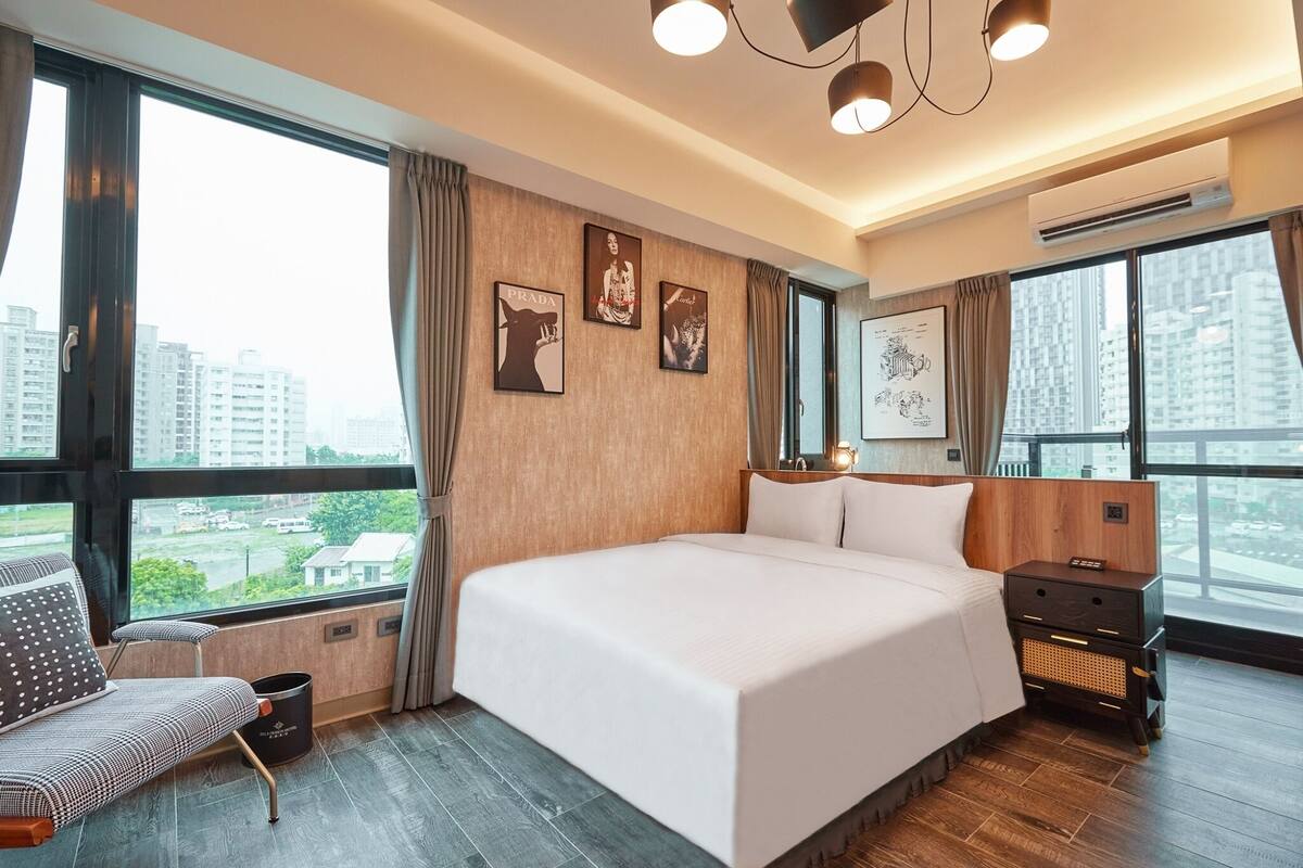 A modern hotel room with a large bed, a bedside table, a chair, and a view of the city through large windows.