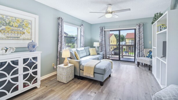 A spacious room with a ceiling fan, a large window with curtains, a sofa, a side table, and a cabinet with glass doors. - Cocoa Beach Vacation Rental - Walk to Pier! (Cocoa Beach)