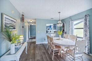 A dining area with a round table, white chairs, and a chandelier. There is a kitchen in the background with blue cabinets. - Cocoa Beach Vacation Rental - Walk to Pier! (Cocoa Beach)