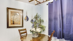 A dining area with a wooden table, chairs, a potted plant with flowers, a framed artwork, and a blue curtain. - Walkable Paradise: Downtown Puyallup Studio! (Puyallup)