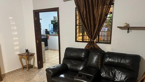 A living room with a black leather sofa, a wooden coffee table, and a small wooden side table. A window with curtains is visible, and a door leads to an outdoor area. - Nanda Parbat Hostal (San Rafael Cedros)